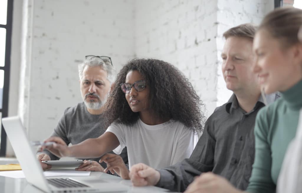 A young black woman wearing glasses and a white top pointing at a laptop screen with a white man on her left side and a white man and white woman on her right side.