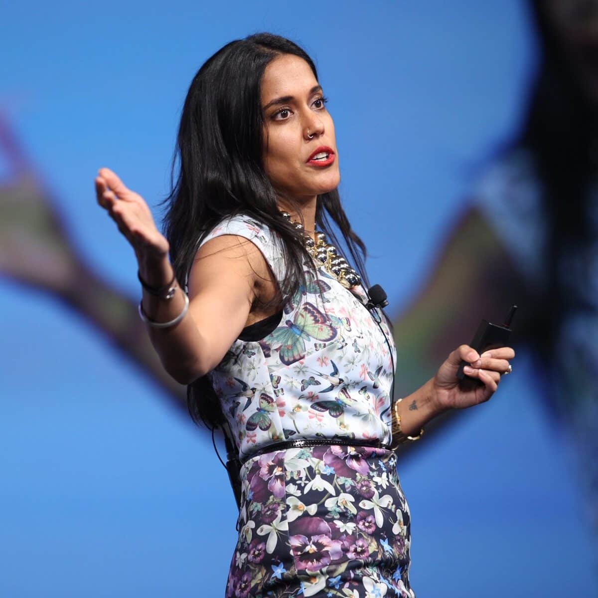 Ritu Bhasin on stage speaking in front of a screen with a wireless remote in one hand and her other arm extended in an explanatory gesture.