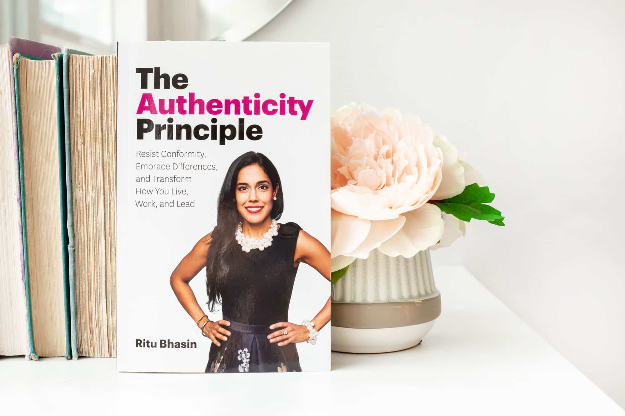 Ritu Bhasin's book The Authenticity Principle sitting on a white table in front of a stack of old books with the pages facing forward and a vase with a large pale pink flower.