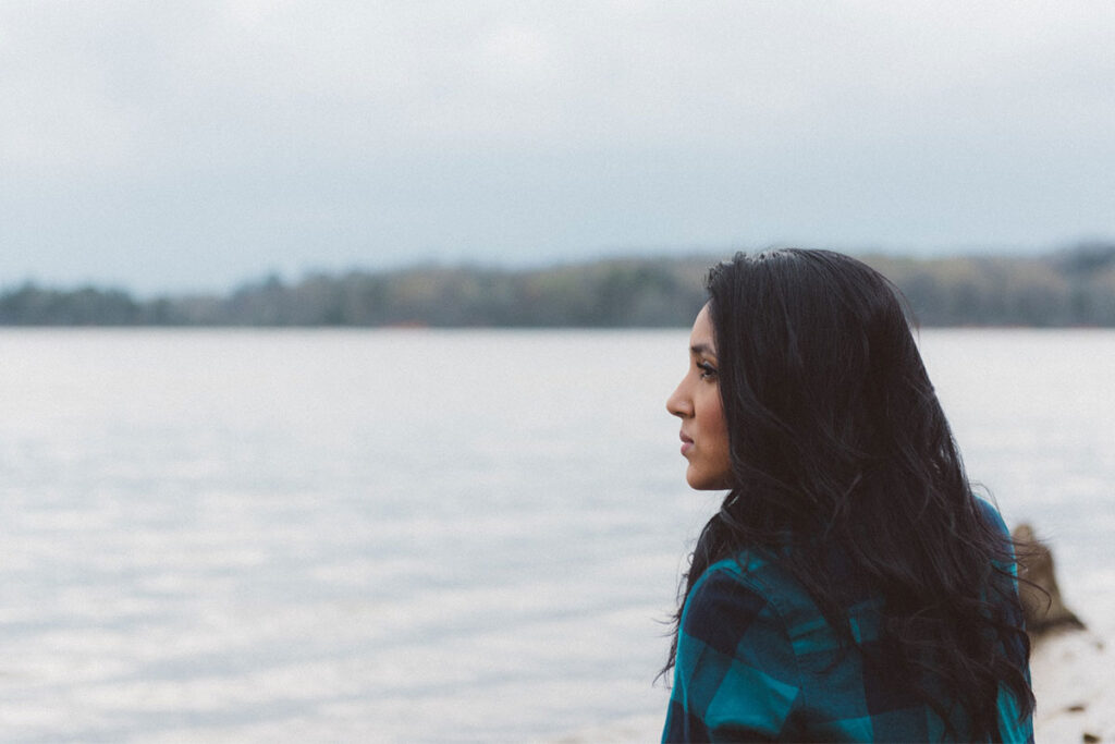A woman with long dark hair wearing a blue plaid shirt looking off to the left side, with a lake and trees in the background