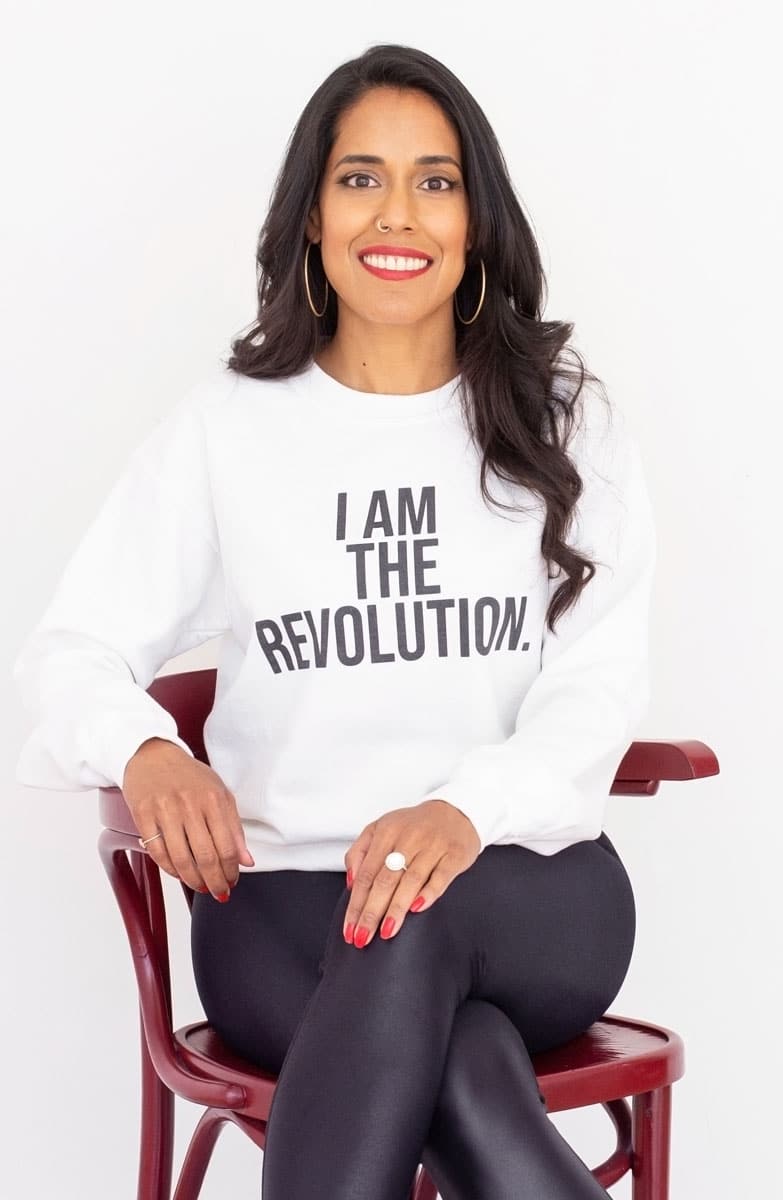 Ritu Bhasin sitting comfortably in a chair, smiling at the camera wearing a white sweatshirt that says "I Am the Revolution"