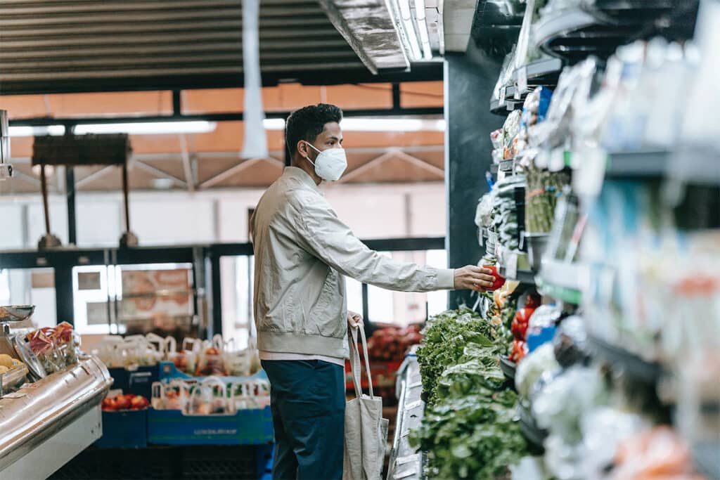 A South Asian man wearing a cream colored jacket, blue pants and a white face mask picking up produce off a grocery store shelf