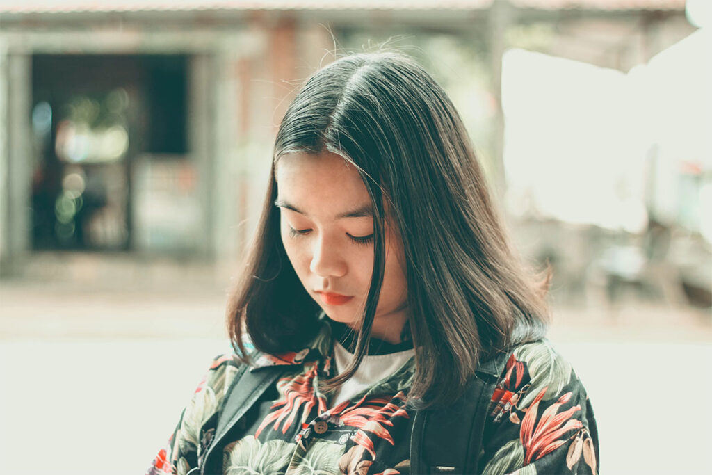 A young East Asian women with black hair wearing a floral top looking down towards the ground.