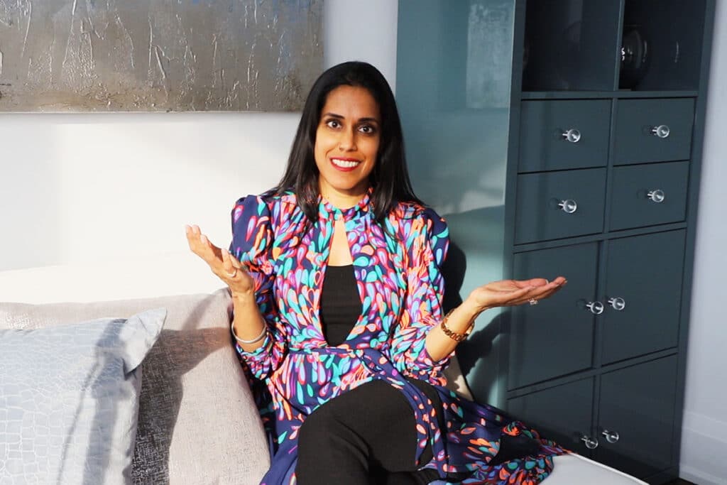 Ritu Bhasin in a multi-colored patterned dress, sitting on a grey sofa with her hands raised slightly.