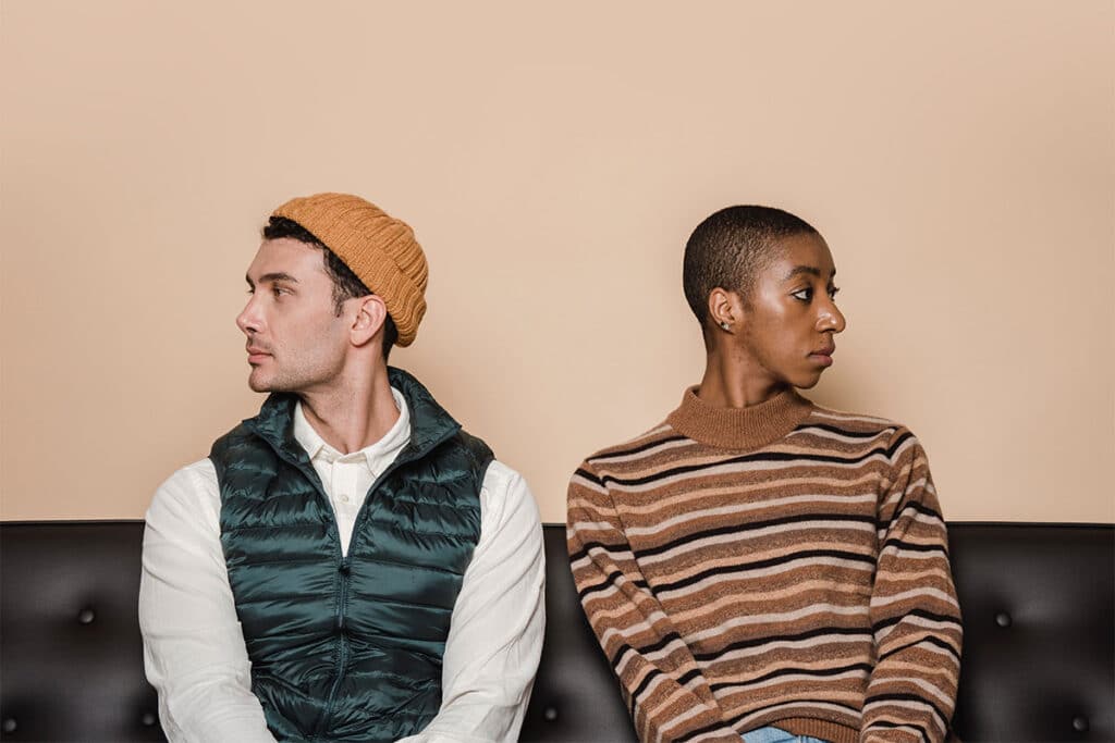 Two people sitting side by side on a black couch looking away from each other. The person on the left is white and is wearing a white shirt and a dark green puffer vest with a yellow hat. The person on the right is black and has a buzz cut, and is wearing a brown, black and grey striped sweater.