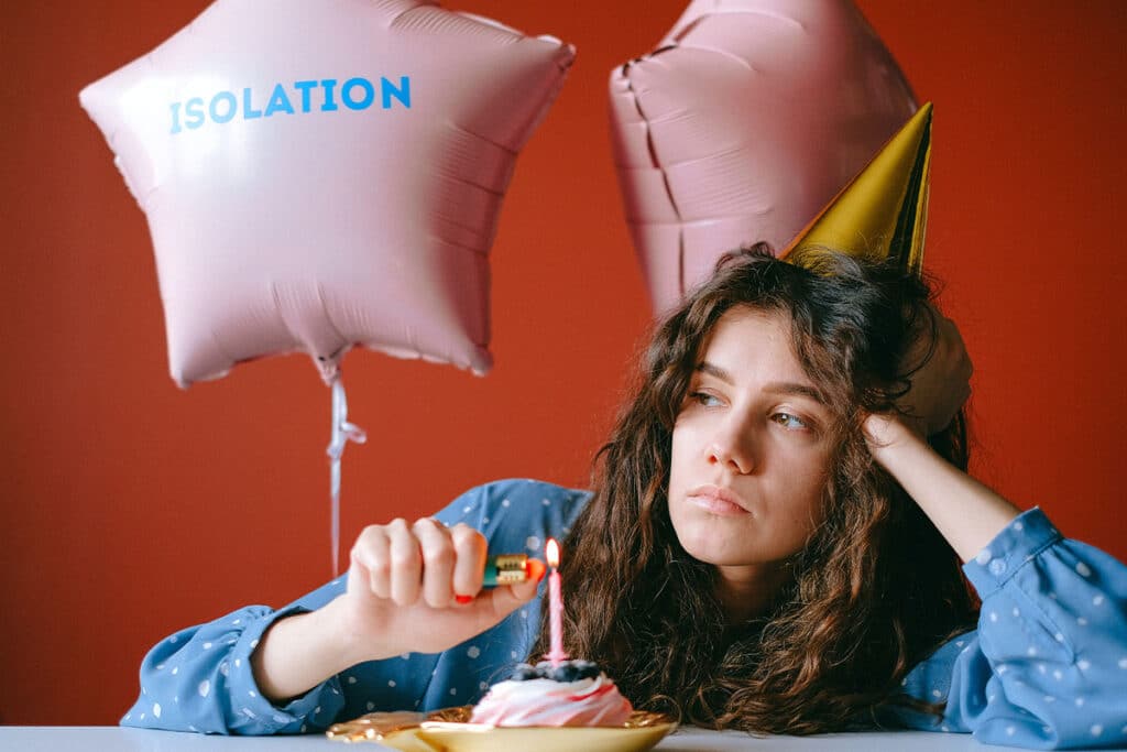 A young woman wearing a yellow party hat looking sad as she lights a candle on a small birthday cake. There is a pink star-shaped balloon behind her that has the word "isolation" printed on it.