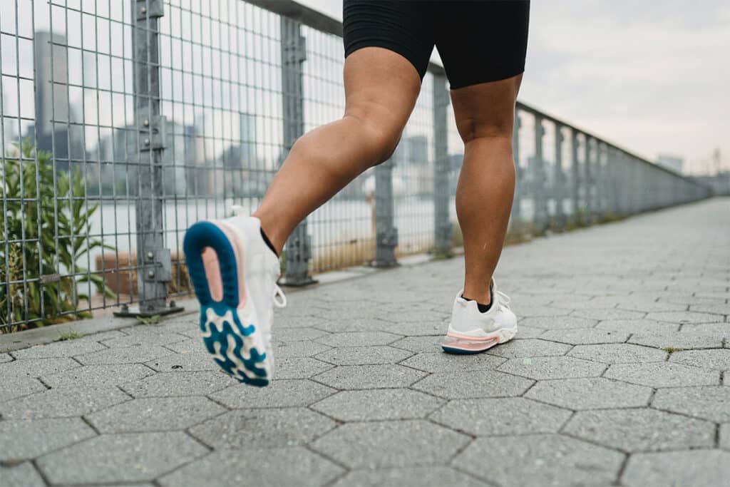 A person jogging on a city bridge with concrete tiles, with only their bottom half of their body in view. They are wearing black running shorts and black socks, with white, pink and blue running shoes.