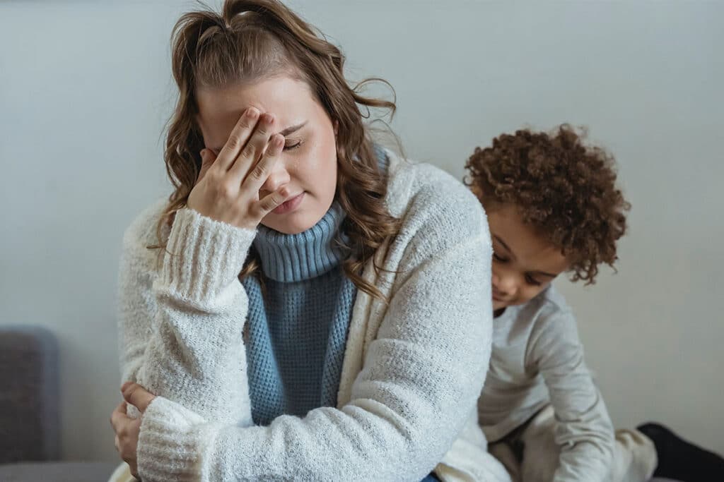 A young woman holding her head in her hand in exasperation with a small child behind her.