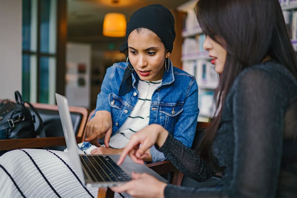 An Asian woman with long dark hair wearing a black long-sleeve top and a Brown woman wearing a black headscarf and blue denim jacket looking at a laptop computer together and pointing at the screen.