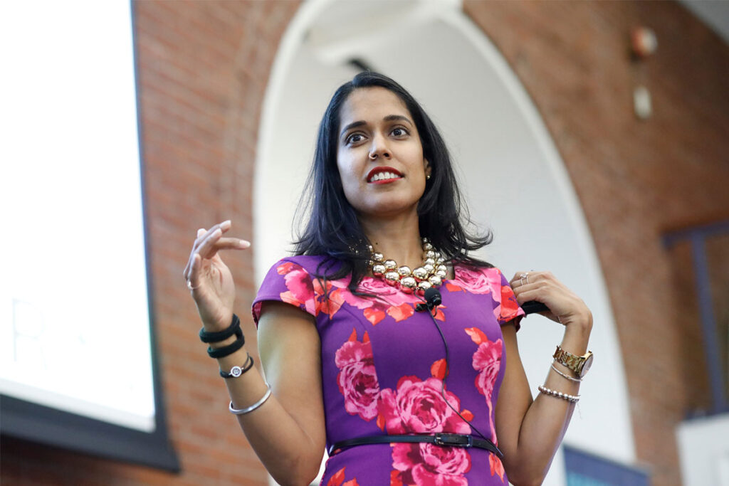 Ritu Bhasin on a stage during a presentation. She is wearing a pink and purple floral dress with a gold necklace.