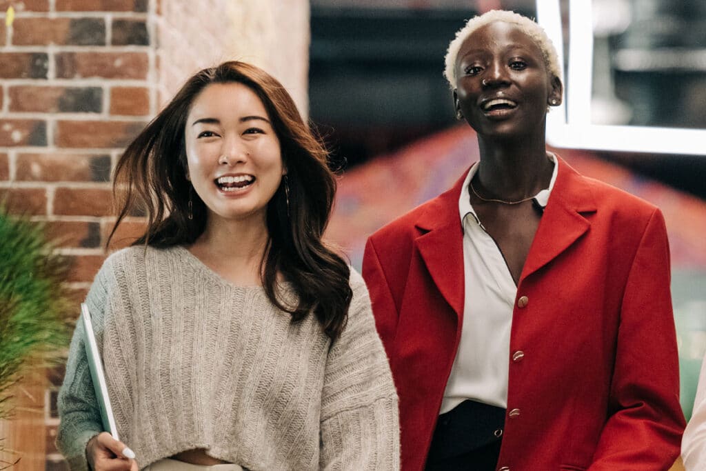 An Asian woman with long dark hair wearing a cream colored sweater and a black woman with short blonde hair wearing a red blazer smiling as they walk