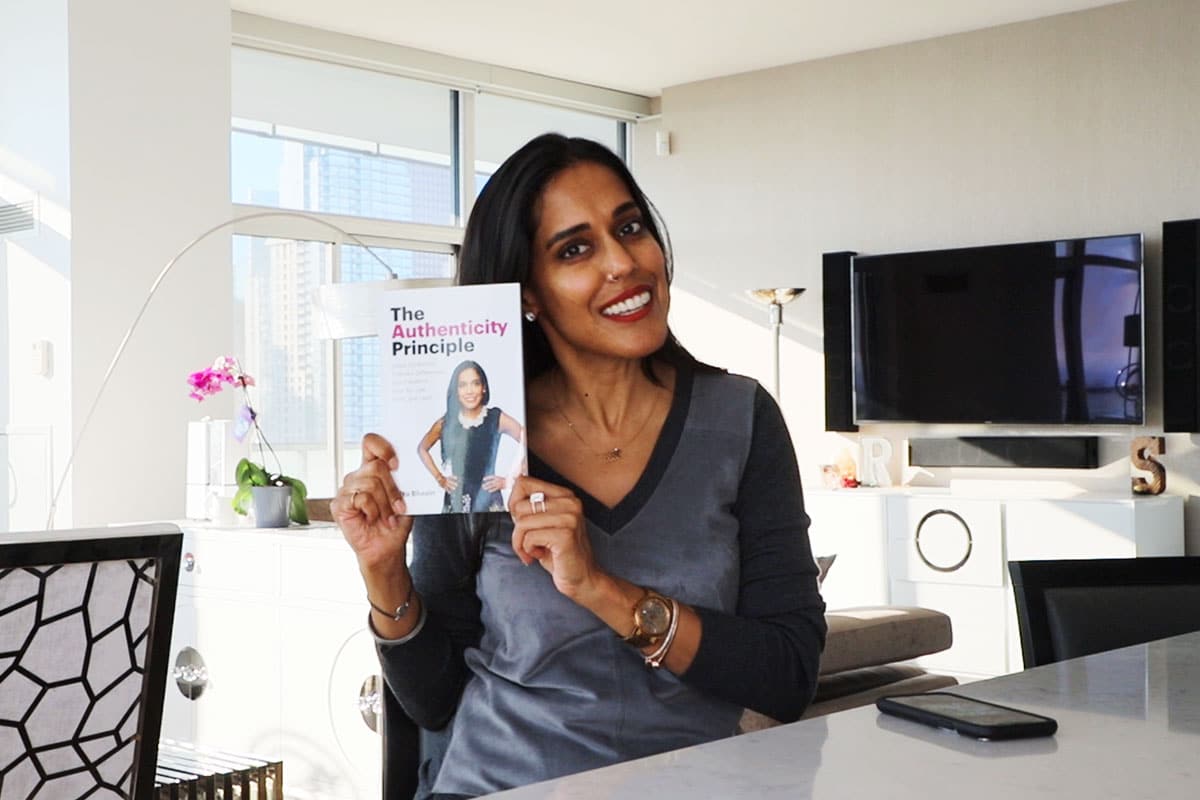 Ritu Bhasin sitting and smiling while holding up a copy of her book, "The Authenticity Principle"