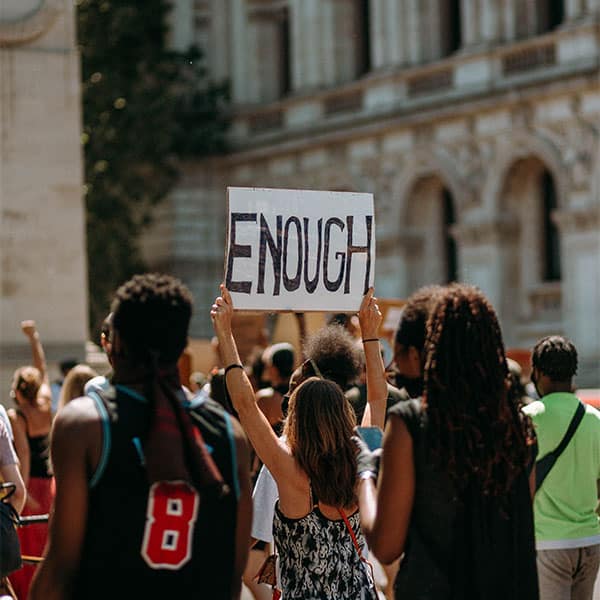 The backs of a group of protestors from various racial backgrounds, with a woman in the middle holding a sign that reads "ENOUGH"