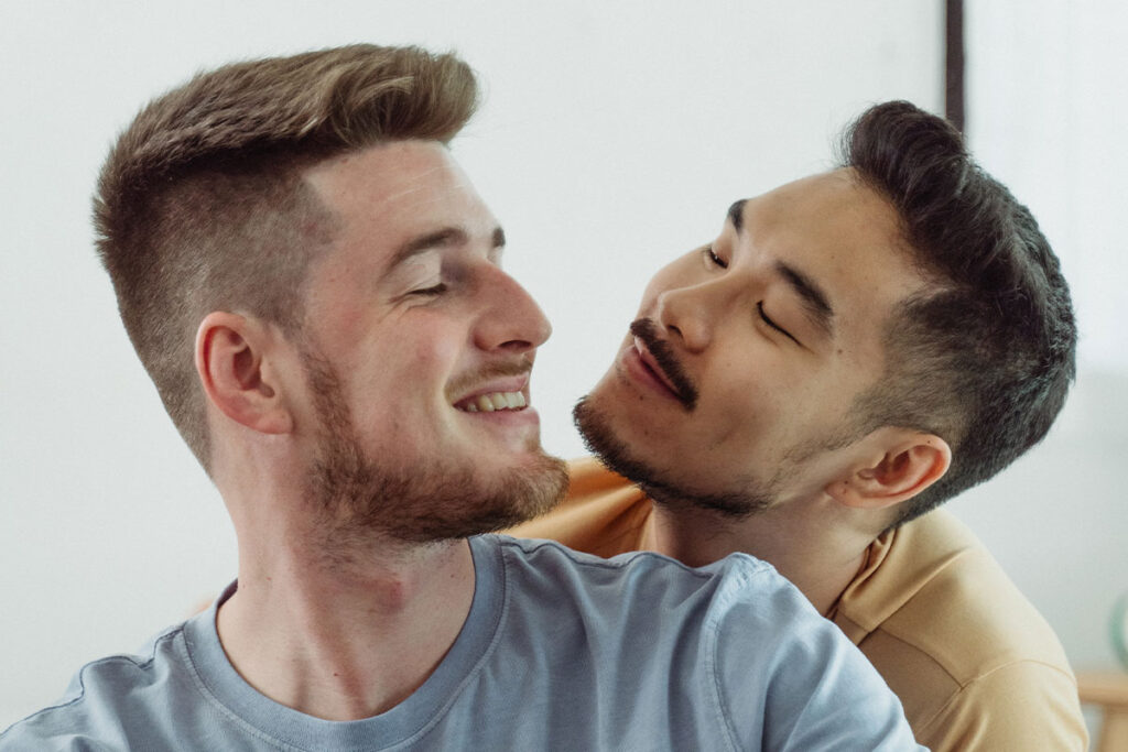 Two men in a relationship smiling and looking at each other. The man on the left is white with blonde hair and beard and is wearing a pale blue t-shirt. The man on the right is East Asian with black hair and beard, wearing a pale yellow t-shirt.