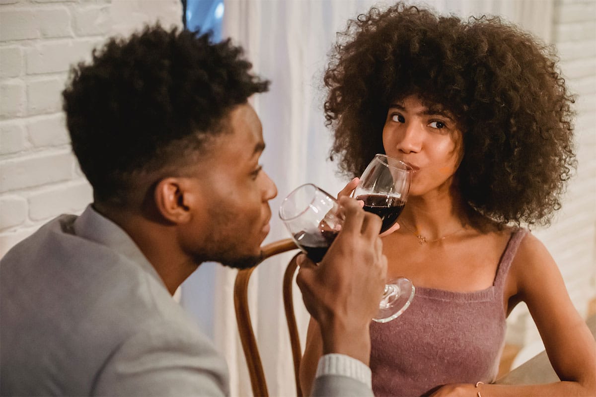 A young black heterosexual couple drinking glasses of wine. The man is on the left side wearing a grey blazer. The woman is on the right and is wearing a mauve tank top, while looking off to the right side.