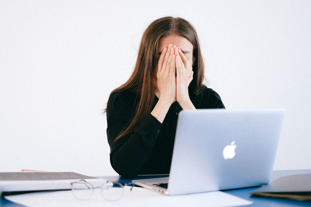 A woman wearing a black shirt with long brown hair sitting at a desk in front of a computer with her head in both hands.