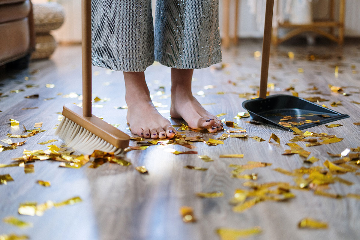 A pair of feet standing on a floor covered in gold confetti, with a broom on the left and a dustpan on the right.