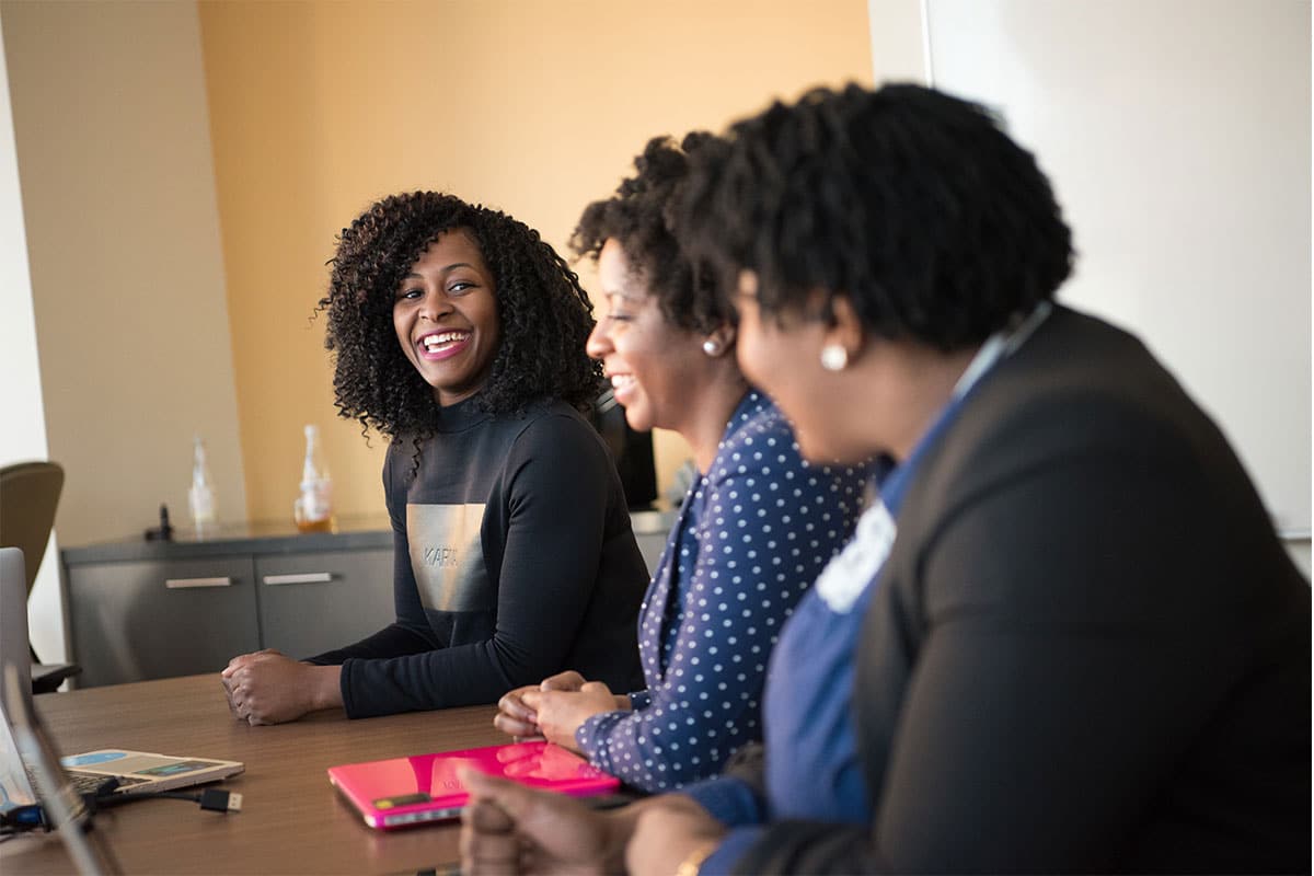 Three black women sitting in a row next to each other at a table, with one of them in focus and smiling at the others.
