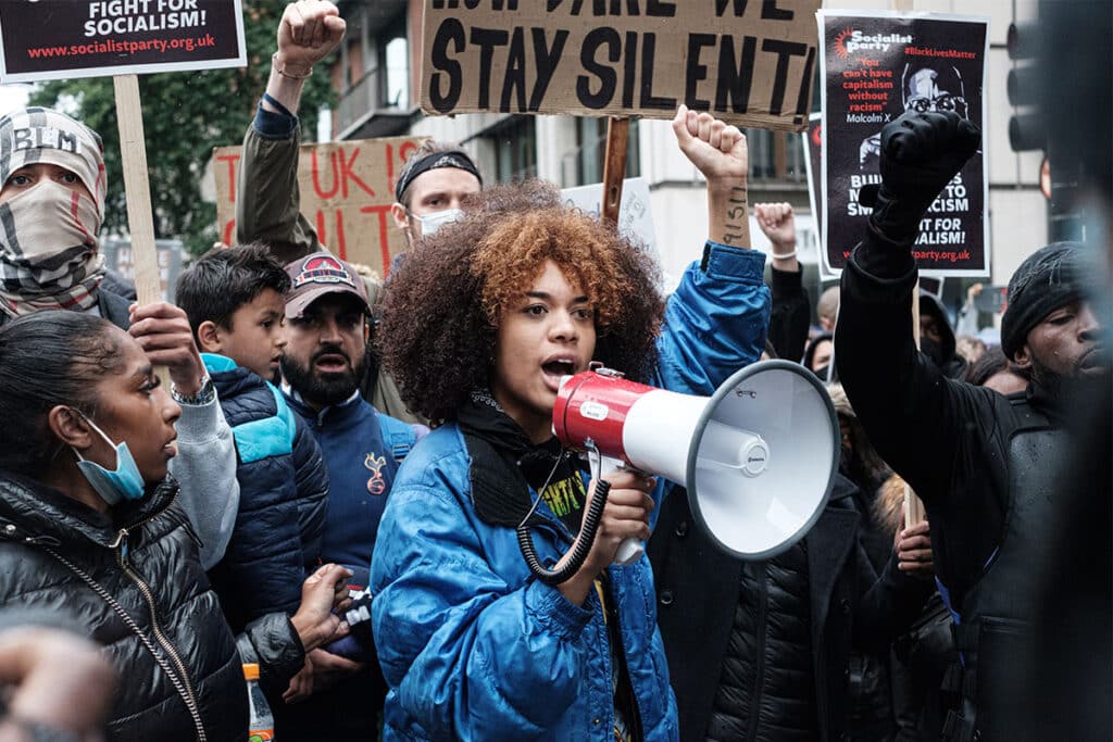 A young black woman wearing a blue jacked and holding a megaphone with her right fist in the air, standing with a group of anti-racism protestors.
