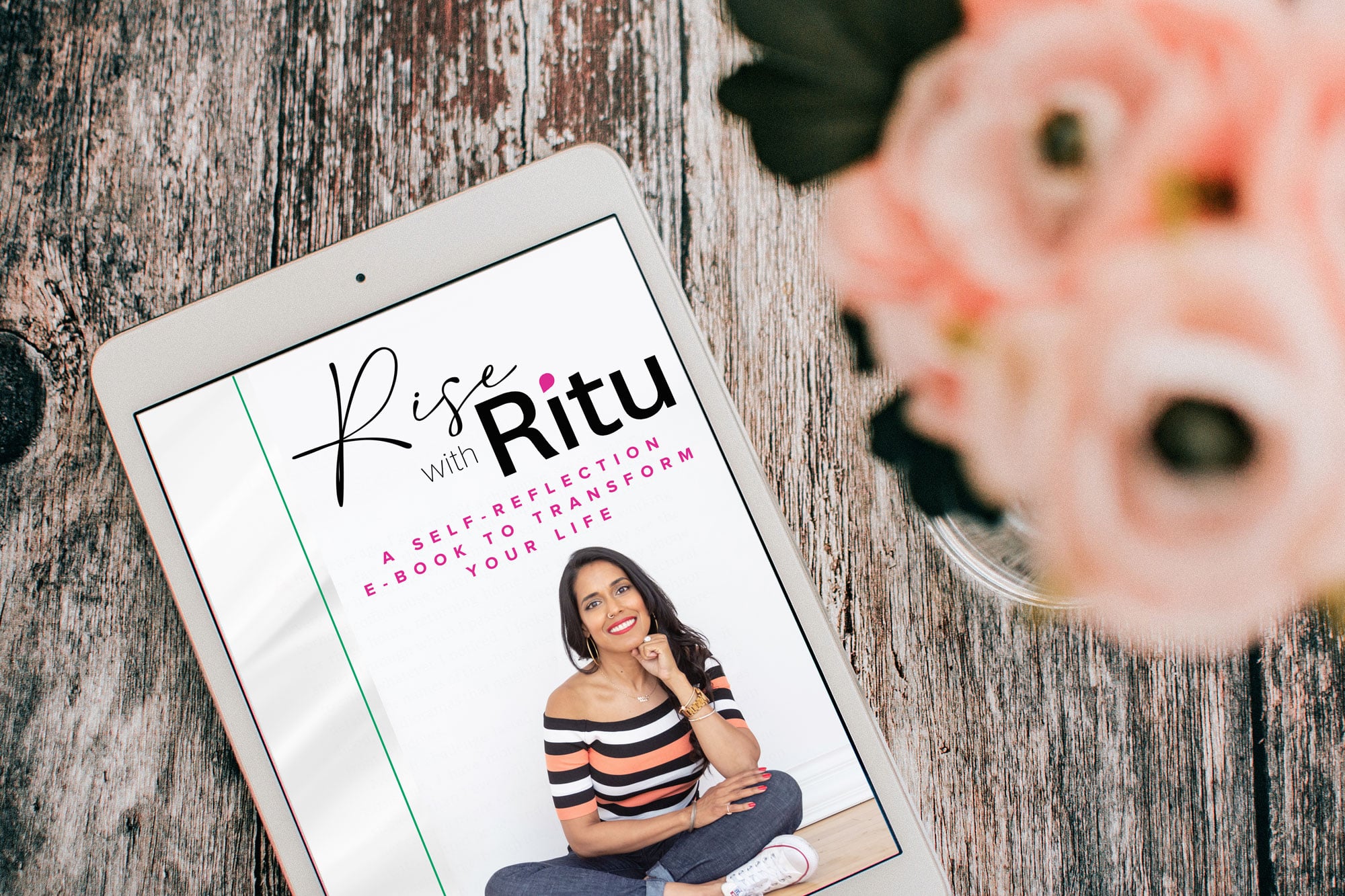 White tablet with Rise with Ritu self-reflection e-book on the screen sits on a wooden table next to a vase filled with pink flowers