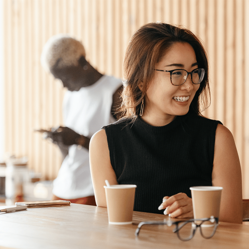 A young Asian woman with glasses sitting at a coffee shop and smiling, with a young Black woman behind her.