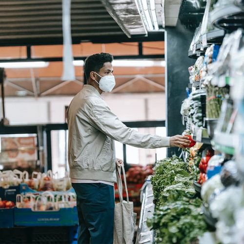 A South Asian man wearing a cream colored jacket, blue pants and a white face mask picking up produce off a grocery store shelf