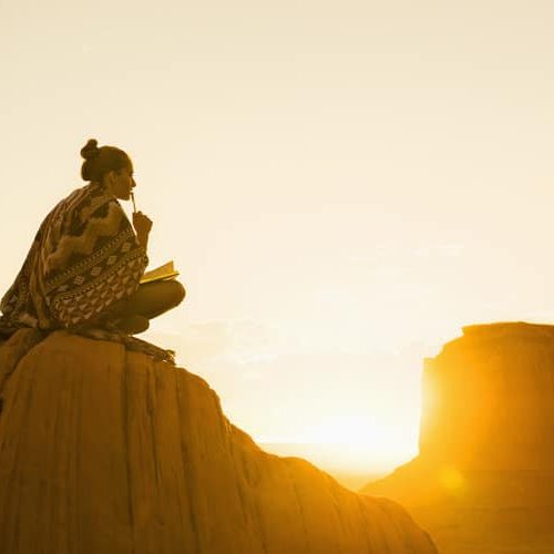 Woman wearing a patterned shawl while sitting with a journal on a hill while the sun rises