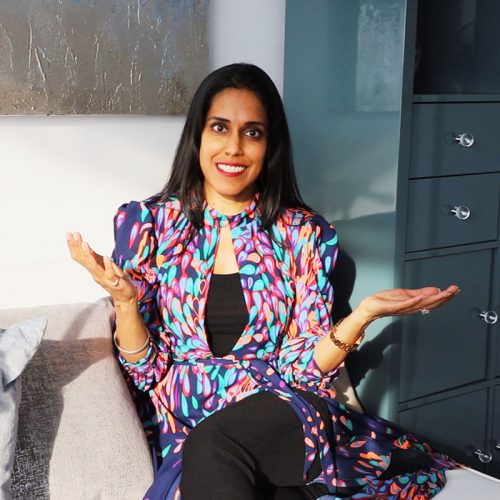 Ritu Bhasin in a multi-colored patterned dress, sitting on a grey sofa with her hands raised slightly.