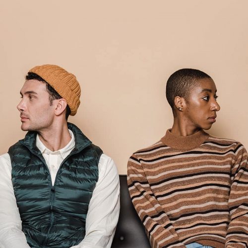 Two people sitting side by side on a black couch looking away from each other. The person on the left is white and is wearing a white shirt and a dark green puffer vest with a yellow hat. The person on the right is black and has a buzz cut, and is wearing a brown, black and grey striped sweater.