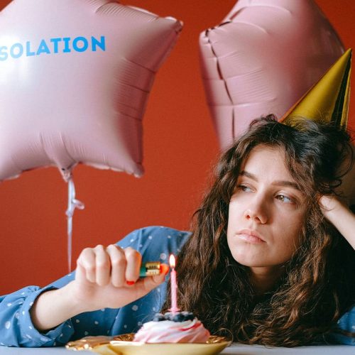 A young woman wearing a yellow party hat looking sad as she lights a candle on a small birthday cake. There is a pink star-shaped balloon behind her that has the word "isolation" printed on it.