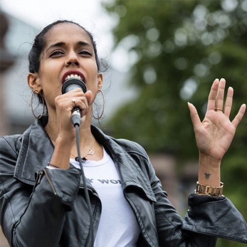 Ritu Bhasin holding a microphone and speaking at a rally outside. She is wearing a white t-shirt and a black leather jacket and her black hair is pulled back. Her left hand is holding the mic and her right hand is raised.