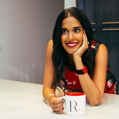 Ritu Bhasin leaning on a white counter holding a white mug with her left hand, and holding her head with her right hand.