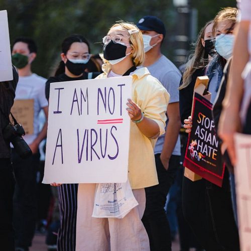A group of protestors outdoors holding up signs. A young East Asian woman is in the middle holding a sign that says "I am not a virus".