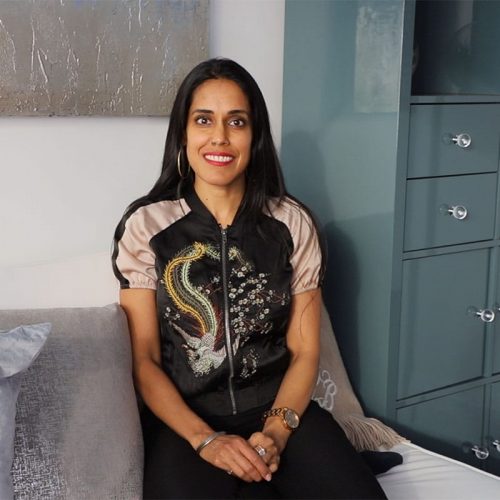 Ritu Bhasin sitting on a grey sofa smiling at the camera. She is wearing a black and grey embroidered top.