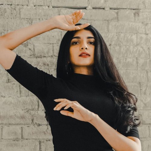 A Brown Woman with long dark hair and wearing a black top posing with one hand placed by her forehead and one in front of her chest