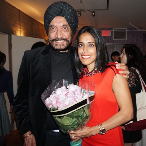 Ritu Bhasin smiling and posting for the camera with her father on her left side. Ritu is wearing a red sleeveless dress and is holding a bouquet of flowers. Her father is wearing a black top and blazer and a black patterned turban.