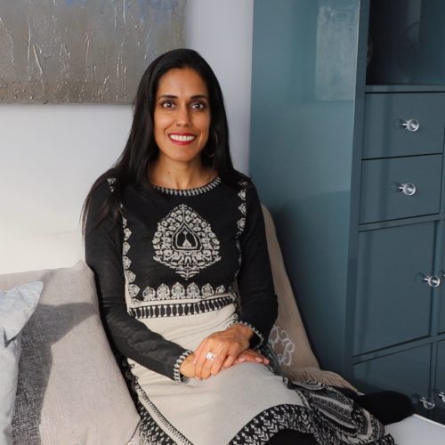 Ritu Bhasin wearing a white and black patterned dress sitting on a grey sofa, facing the camera with her hands placed in her lap.