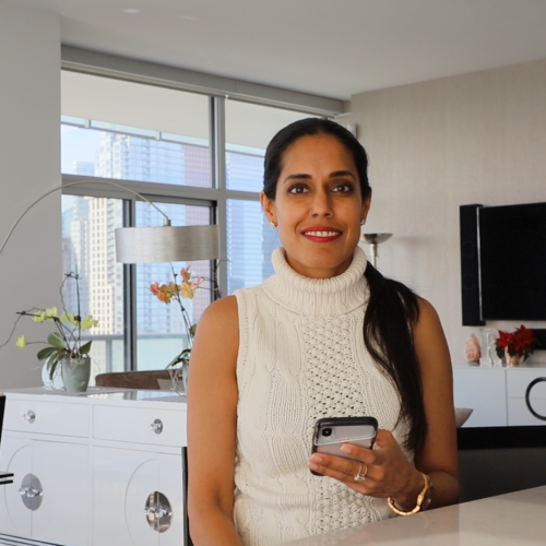 Ritu Bhasin is sitting in a living room wearing a white sleeveless top holding her phone in her right hand, smiling at the camera. Her black hair is tied back into a ponytail and draped over her right shoulder.