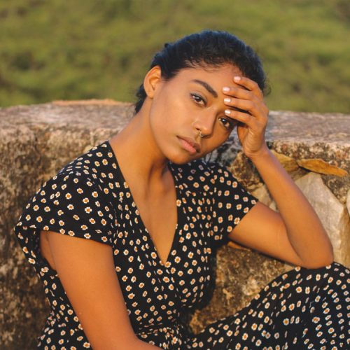 A young woman of color sitting and posing with her head in her right hand, wearing a floral printed jumpsuit.