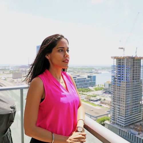 Ritu Bhasin wearking a bright pink sleeveless top with her hair down, standing on an apartment building balcony in daylight looking off to the right side