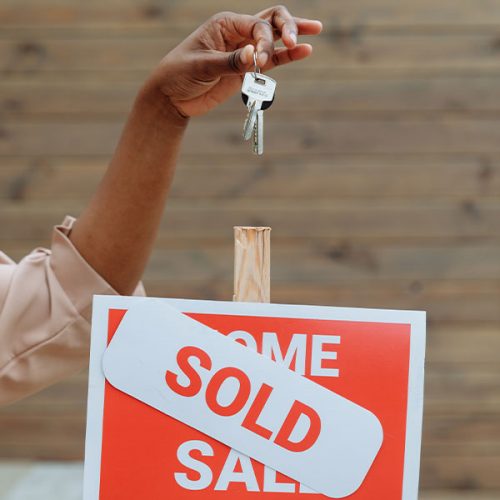 A hand holding up a set of keys above a red and white "Home for Sale" sign with a large "SOLD" sticker across it