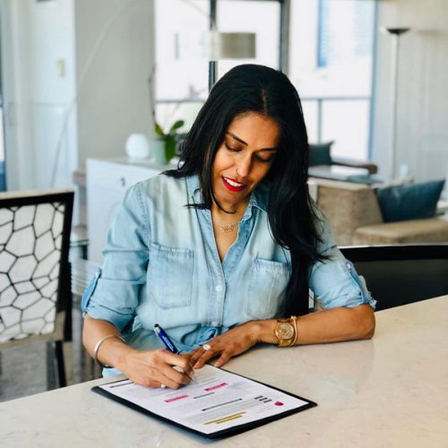 Ritu Bhasin wearing a blue button down shirt sitting at a kitchen counter looking down at a worksheet as she fills it in with a pen.