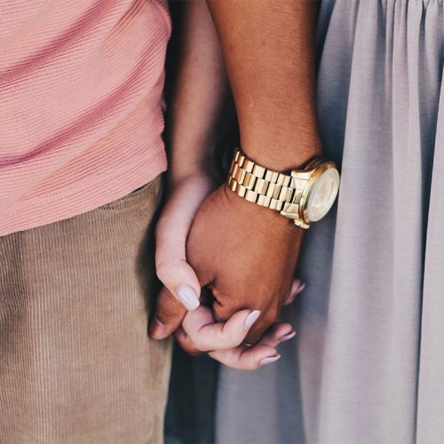 Two people holding hands, one person with a dark brown skin tone wearing a gold wrist watch, and the other person with a paler skin tone wearing grey nail polish