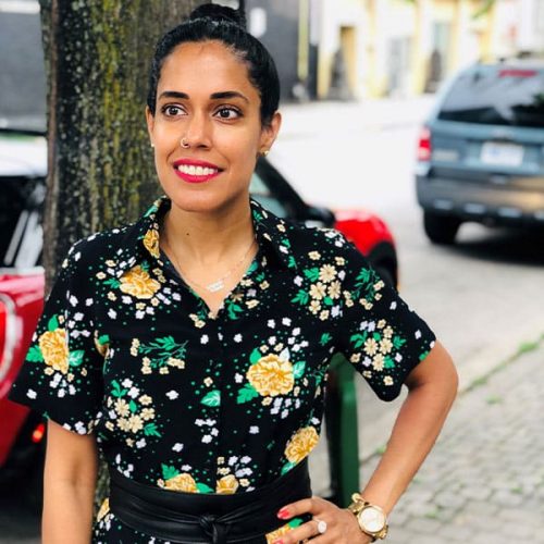 Ritu Bhasin standing on a sidewalk with one hand on her hip, smiling and looking off to her left side. Her black hair is pulled up into a bun and she is wearing a black and yellow floral print dress.