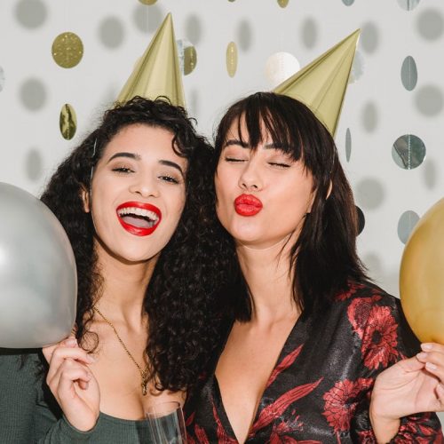 Two young women holding balloons and wearing gold party hats smiling