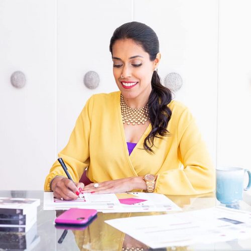 Ritu Bhasin wearing a mustard yellow long sleeve top and her black hair in a pony tail sitting at a desk, looking down at a worksheet while holding a pen.