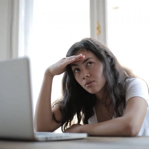 An Asian woman with long dark hair sitting at a desk in front of a computer with her head in her left hand looking off to the right side and looking exasperated.