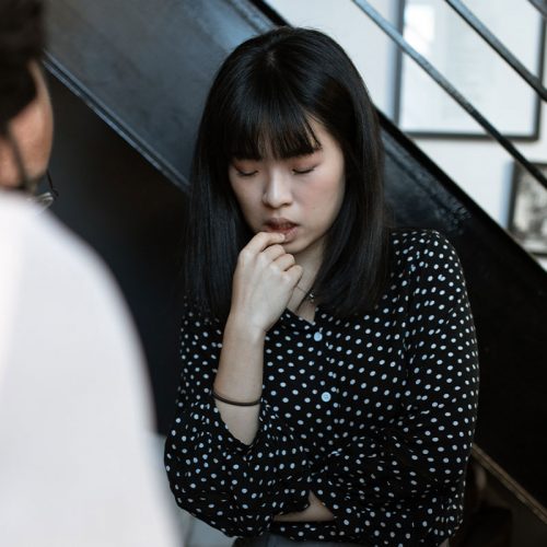A young Asian woman looking nervous as she looks down and her left hand raised to her mouth.