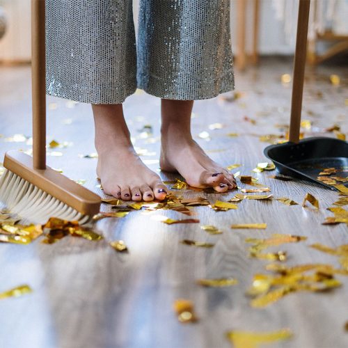 A pair of feet standing on a floor covered in gold confetti, with a broom on the left and a dustpan on the right.