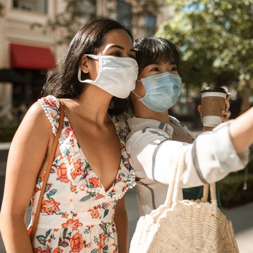 Two young women of color wearing masks and posing for a selfie outdoors.